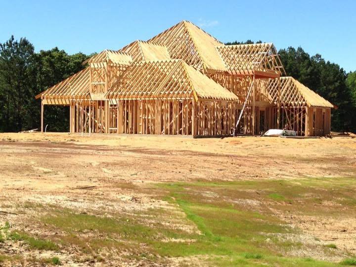 Wood-framed new home with the roof trusses set, standing on a cleared lot under a clear blue sky