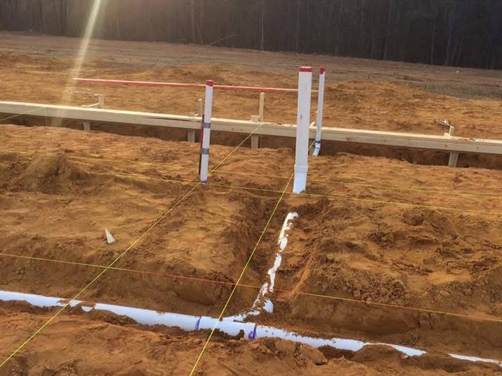 Foundation footing trenches dug in red clay with string lines, wood forms and white plumbing pipes stubbed up, sun setting behind the trees
