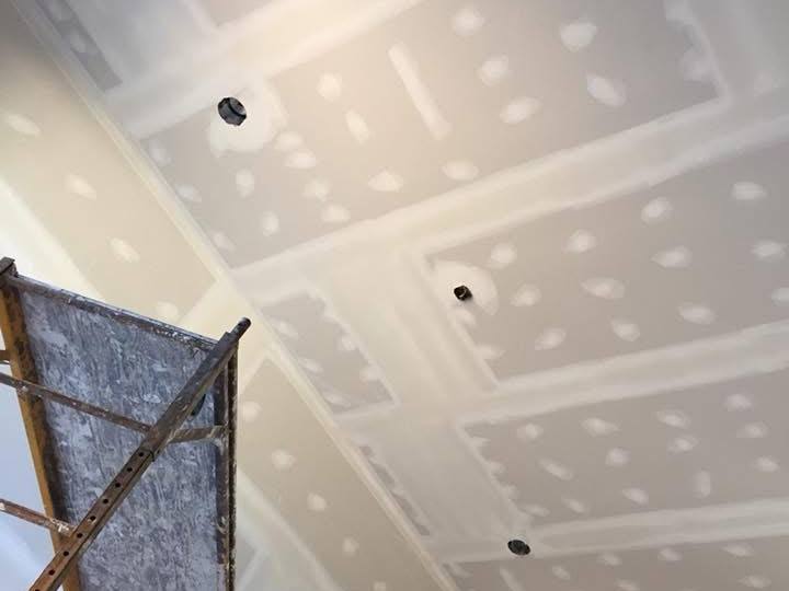 Ceiling drywall with taped and mudded seams and screw spots being finished, with a rolling scaffold in the corner