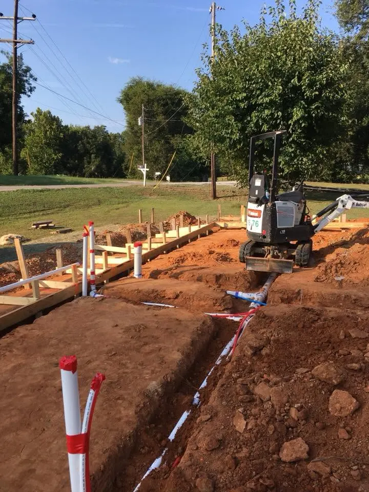 Trench with rough plumbing lines in red clay, a small excavator in the background