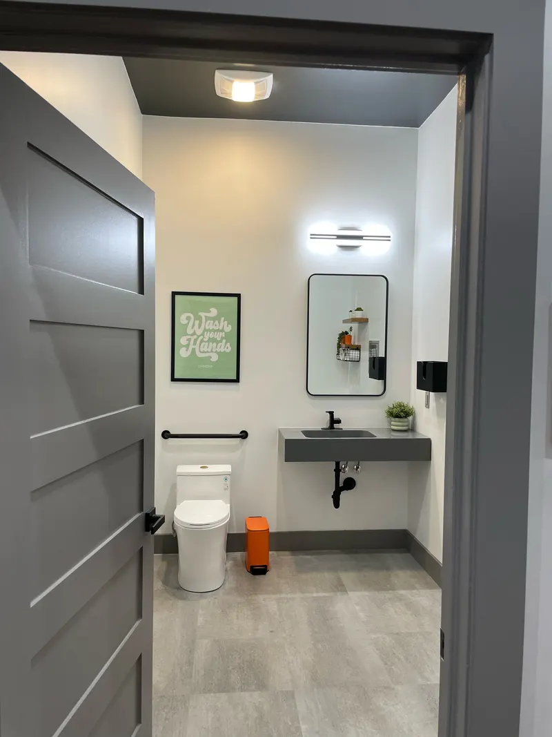 Bathroom with gray floating sink, round mirror, light bar, toilet and grab bar, seen through a doorway