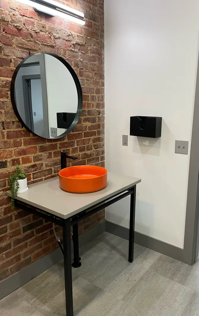 Orange vessel sink on a gray counter with black metal legs against an exposed brick wall, with a round mirror and light bar