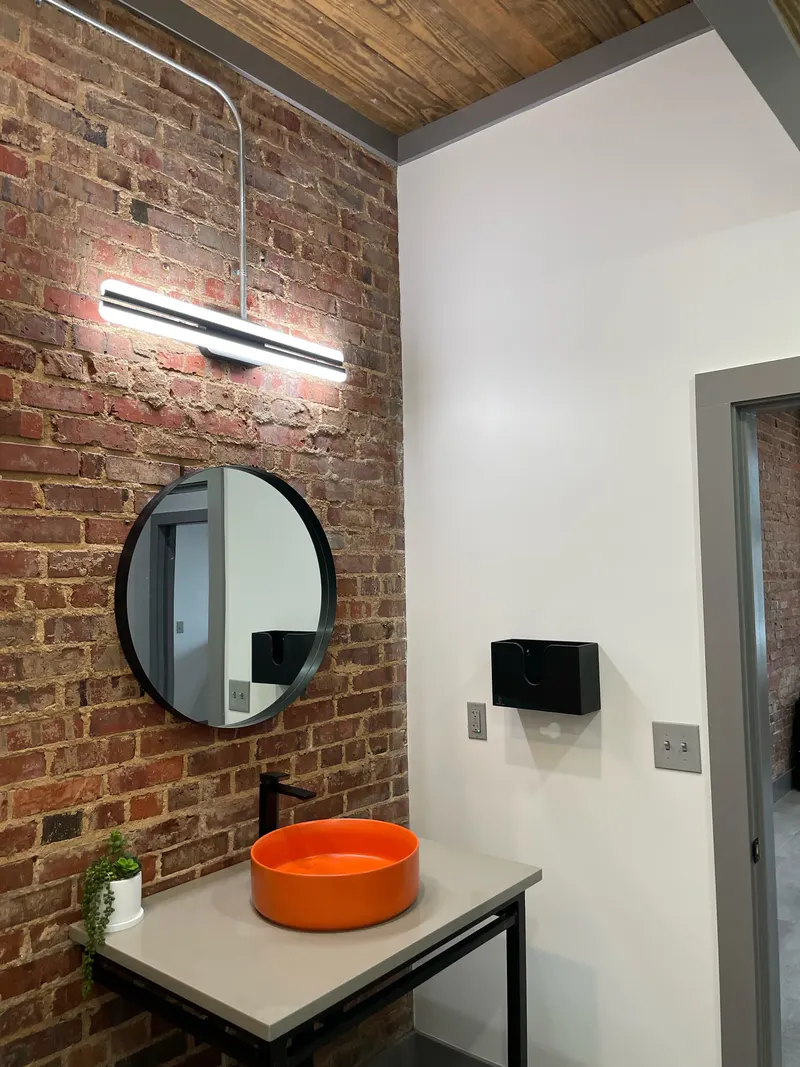 Exposed brick wall with round mirror, light bar and orange vessel sink, under a wood plank ceiling