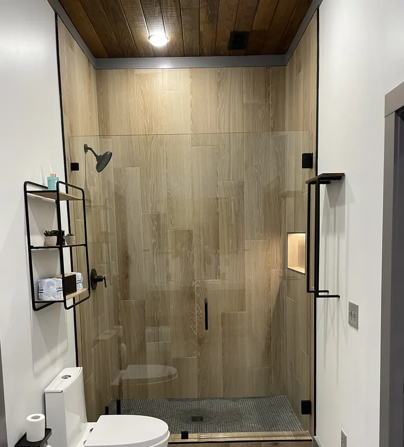Walk-in shower with wood-look tile, glass door and black fixtures, with a wall shelf and toilet beside it