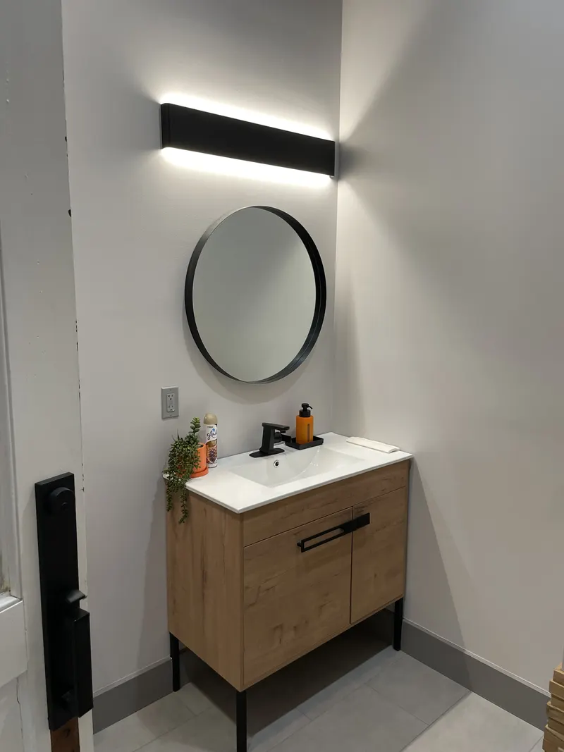 Wood vanity with white top and black faucet under a round mirror and a black light bar