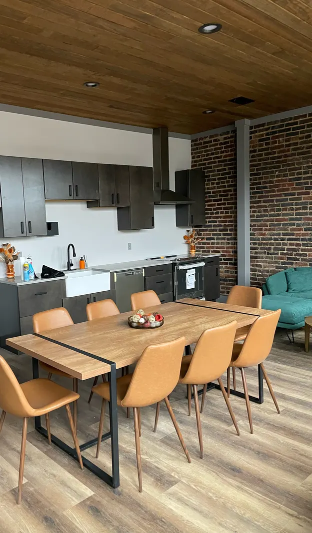 Open kitchen with dark cabinets, farmhouse sink and range hood, a wood plank ceiling, exposed brick and a wood dining table with tan chairs