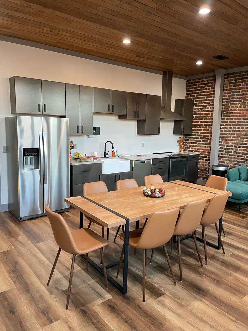Kitchen with dark cabinets, stainless refrigerator and range, wood plank ceiling, brick walls and a long dining table