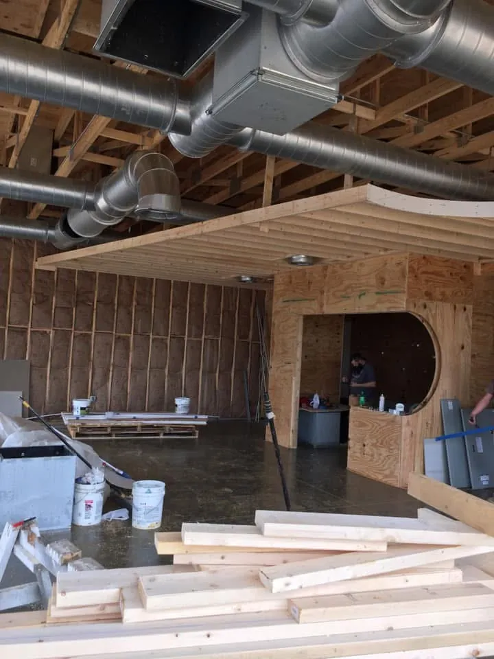Interior under construction with exposed roof framing, spiral HVAC ductwork, a framed curved soffit and a stack of lumber