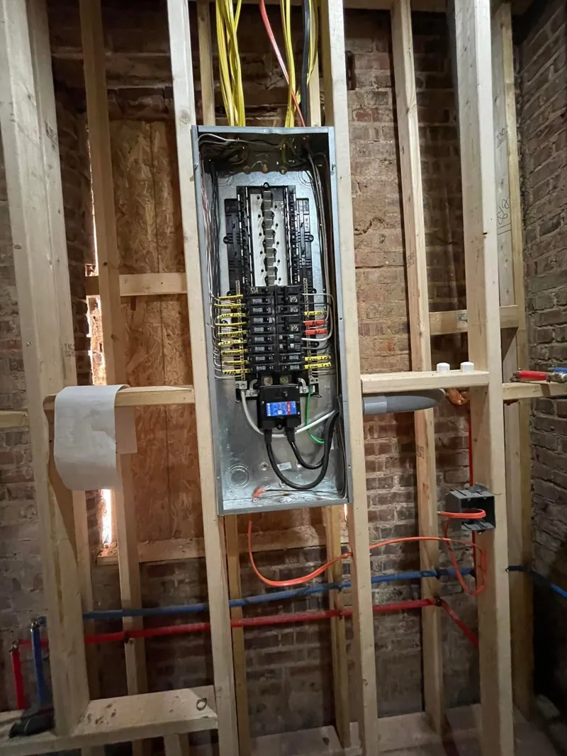 Open electrical panel with labeled breakers mounted between new wood studs, with wiring run overhead and exposed brick behind