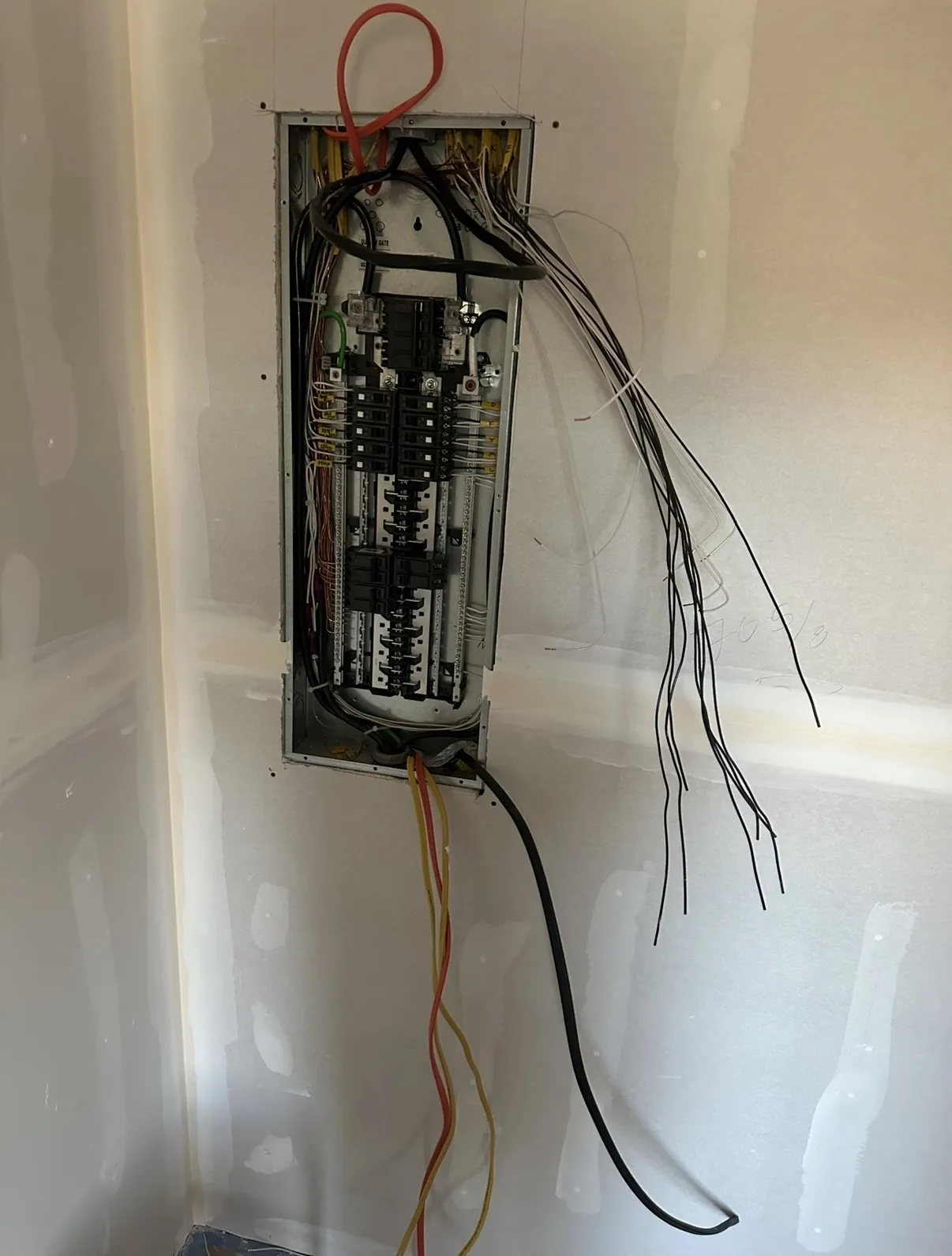 Open breaker panel recessed in unfinished drywall, with breakers installed and bundles of wiring hanging from the sides
