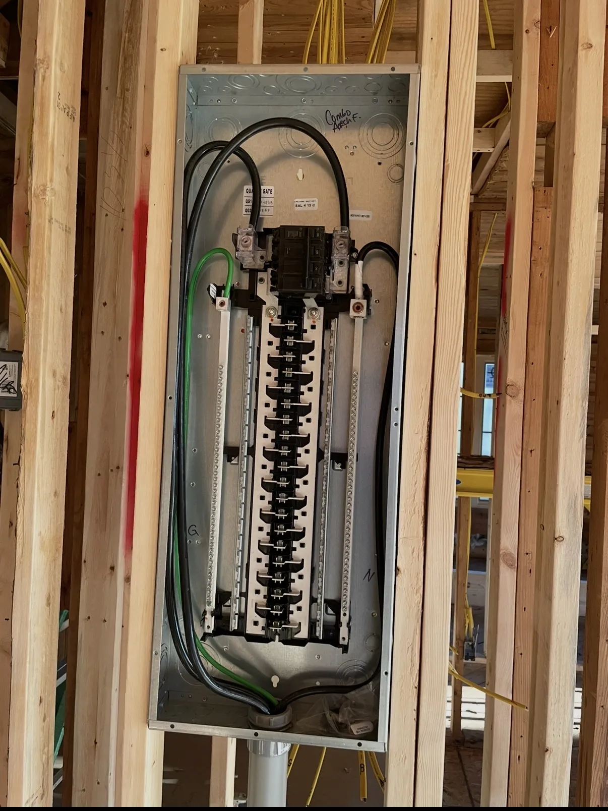 New electrical panel box mounted between wood studs, with a main breaker, service conductors and a handwritten label at the top
