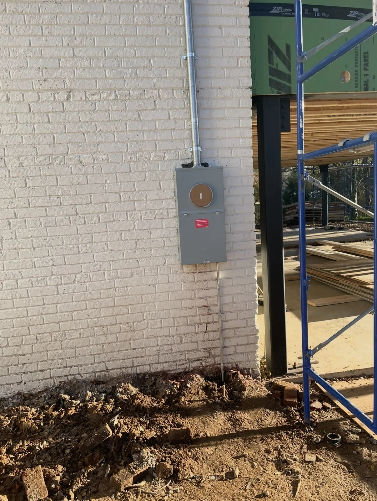 Gray metal meter box mounted on a white brick wall, with conduit above and below and construction debris on the ground