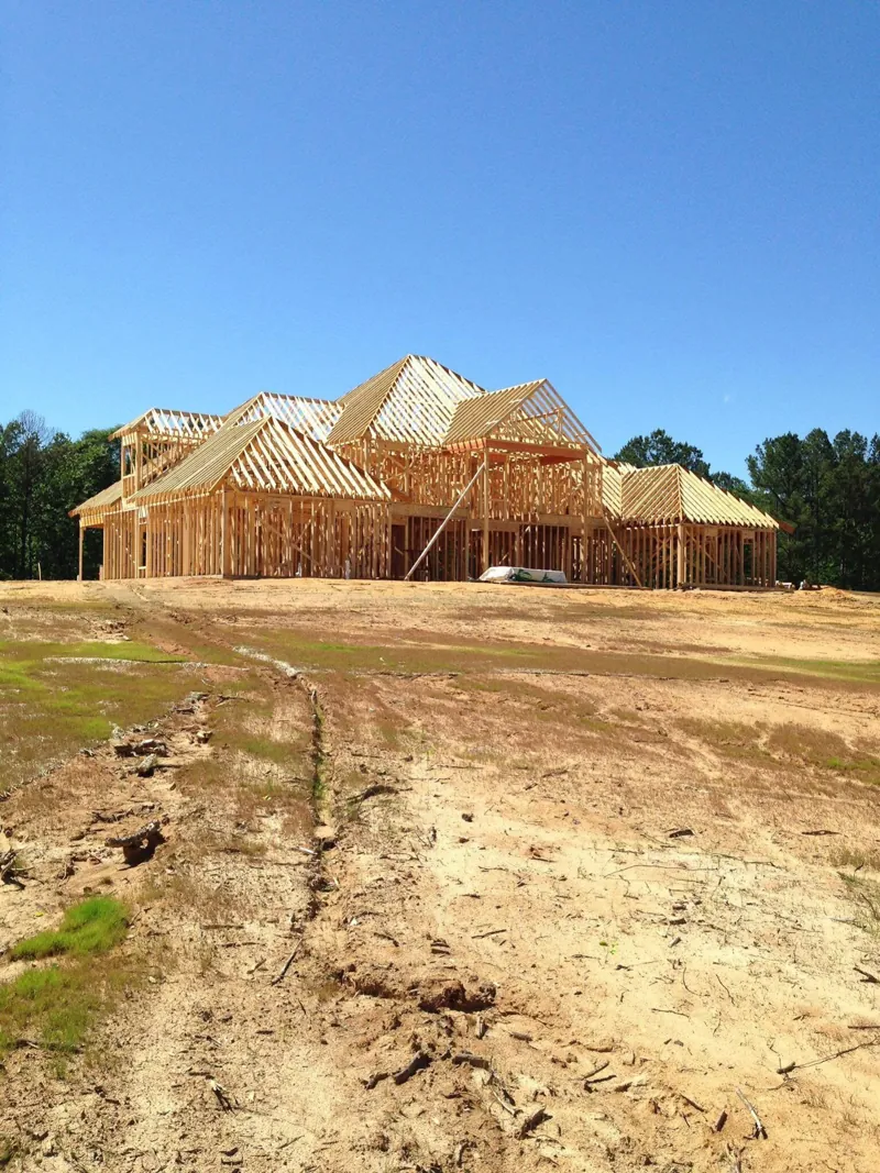 Large house framed with wood walls and roof trusses on a graded hilltop under a blue sky