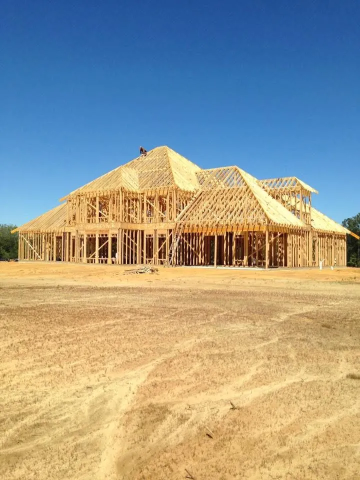 Two-story wood-framed house with a complex roofline across a graded dirt lot, a worker on the roof peak