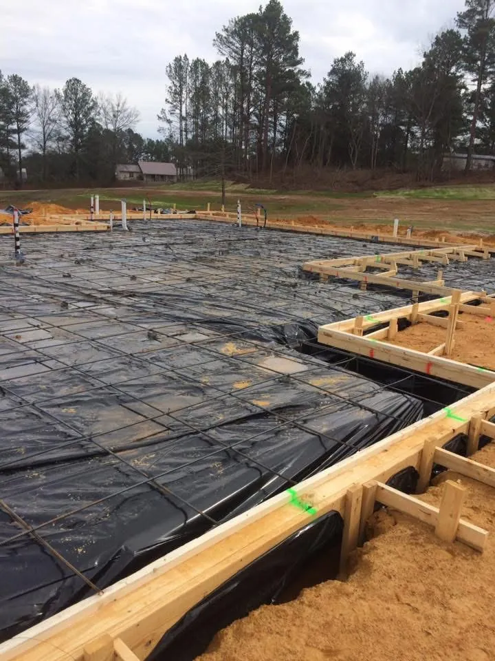 Foundation forms with black vapor barrier and rebar laid out, with woods behind the lot