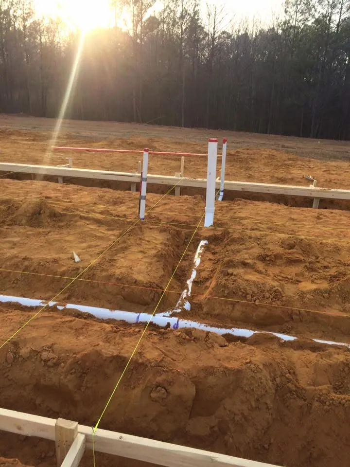 Under-slab plumbing pipes in trenches with yellow layout strings, the sun low over the trees