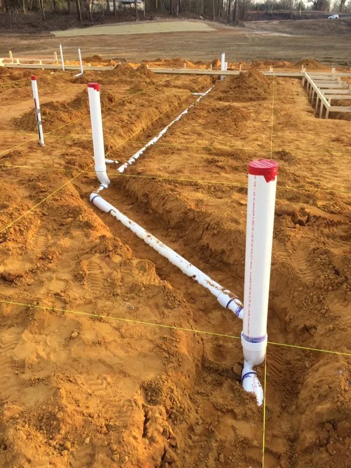 White PVC drain lines and capped stub-ups in trenches of red clay, with layout strings across the pad