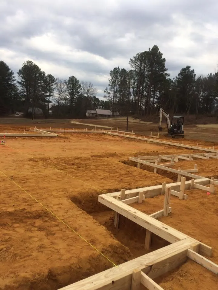 L-shaped wood foundation forms on a cleared lot, with an excavator and trees in the background