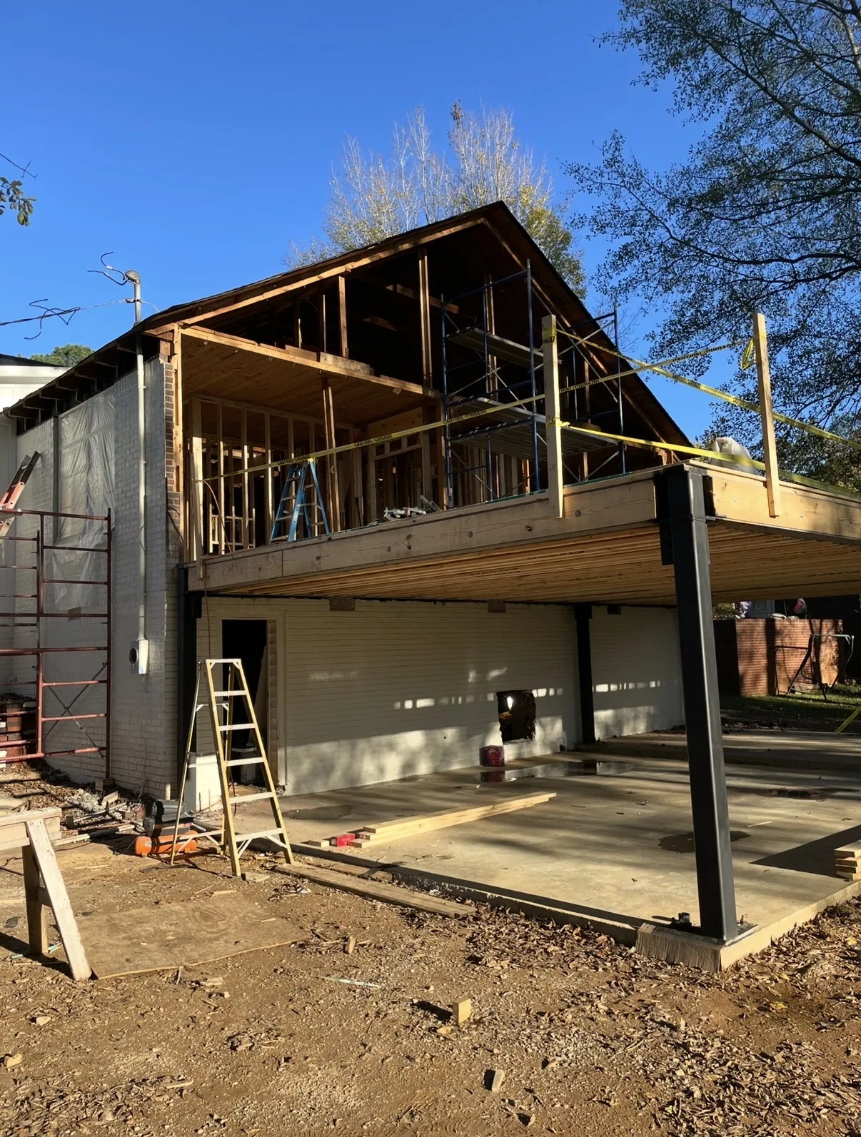 Two-story house addition on a white brick home with steel beams, an open lower patio and scaffolding on the upper deck