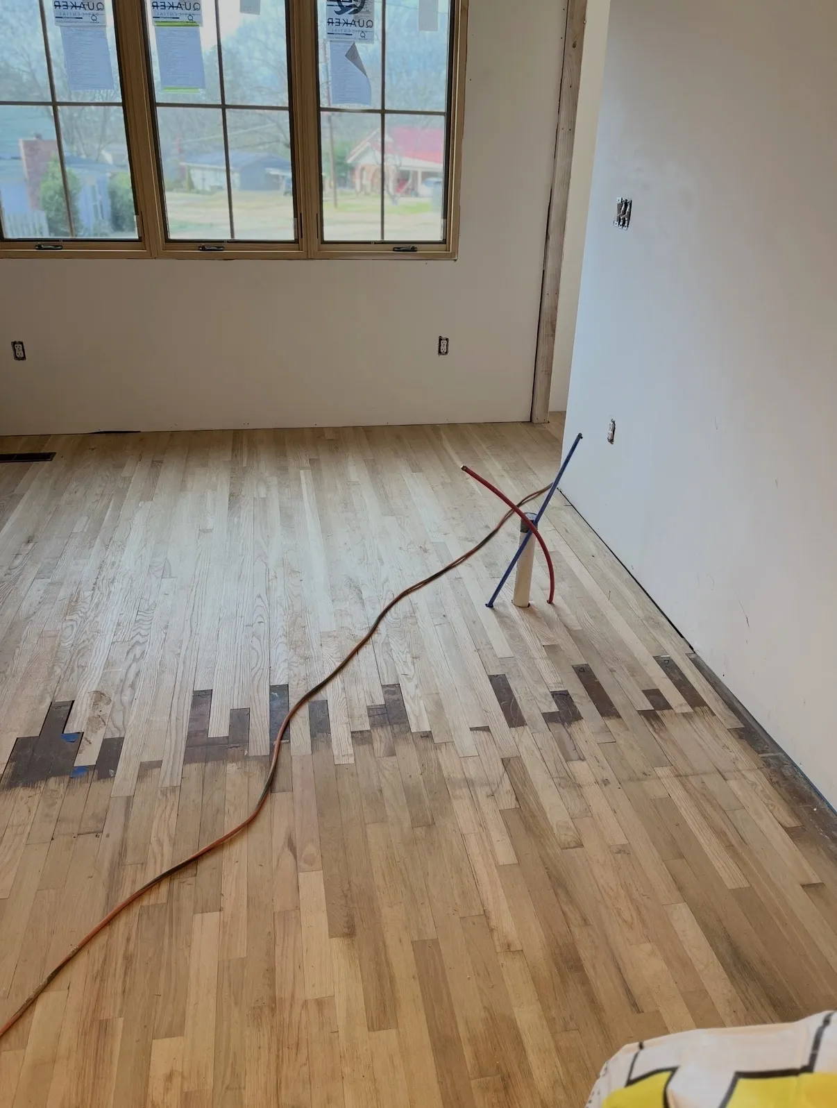 Light oak hardwood flooring mid-install meeting older darker boards, with PVC and red and blue PEX plumbing stubs rising from the floor