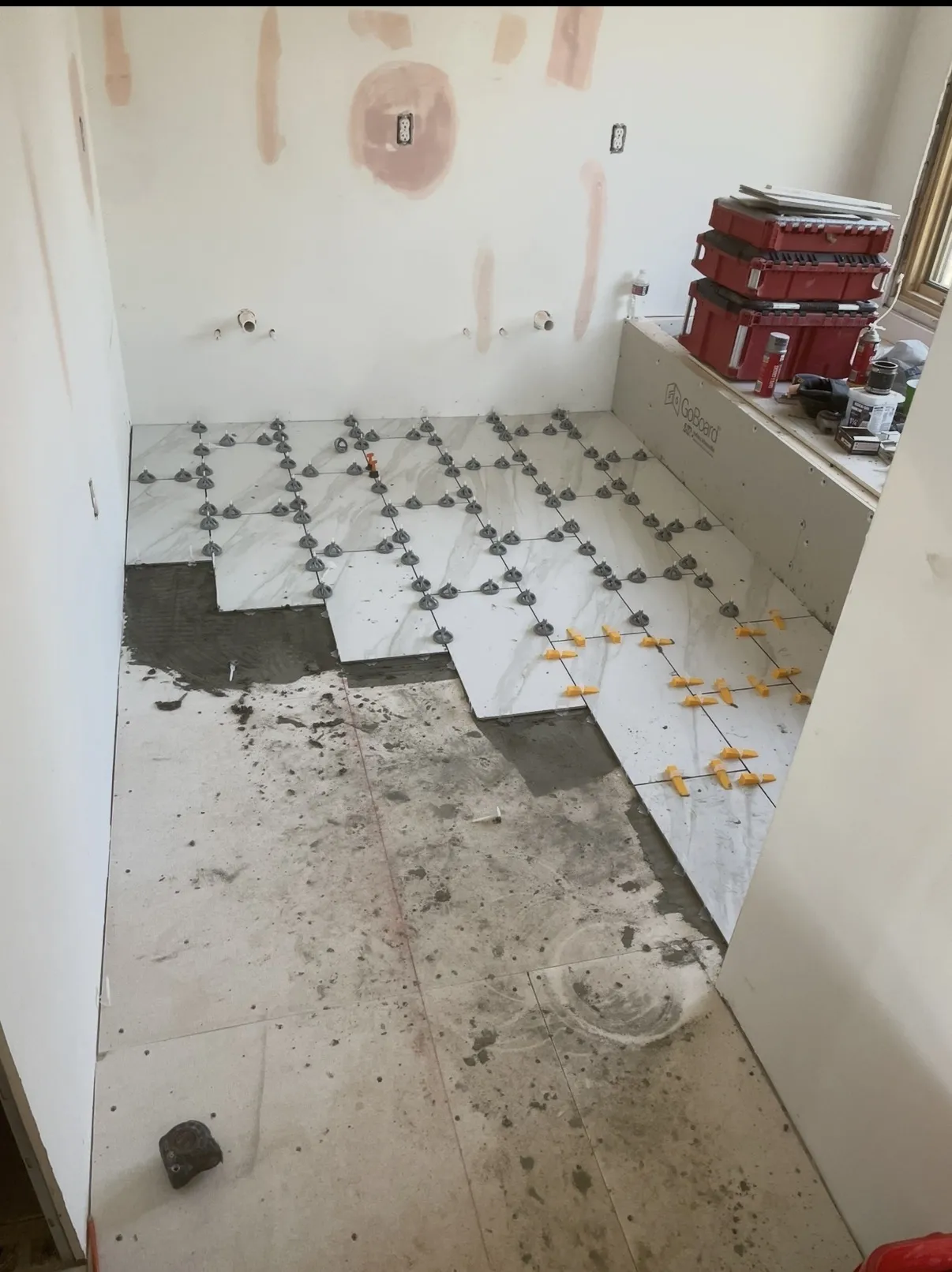 Large white marble-look floor tiles being set with gray leveling clips and orange spacers, plumbing stubs on the back wall